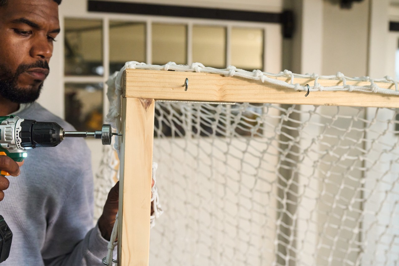 A man uses a cordless screwdriver to attach screw hooks to the struts of the goal