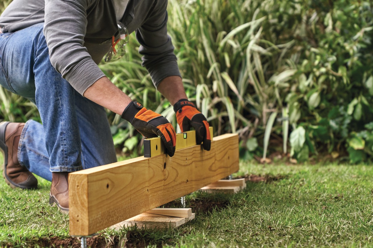 A floor joist is positioned in place over the pile holes and rests on blocks, as someone checks it using a spirit level