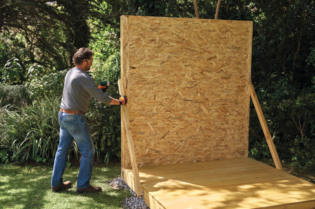 A man mounts a pre-assembled wall onto a wooden DIY shed base using temporary supports