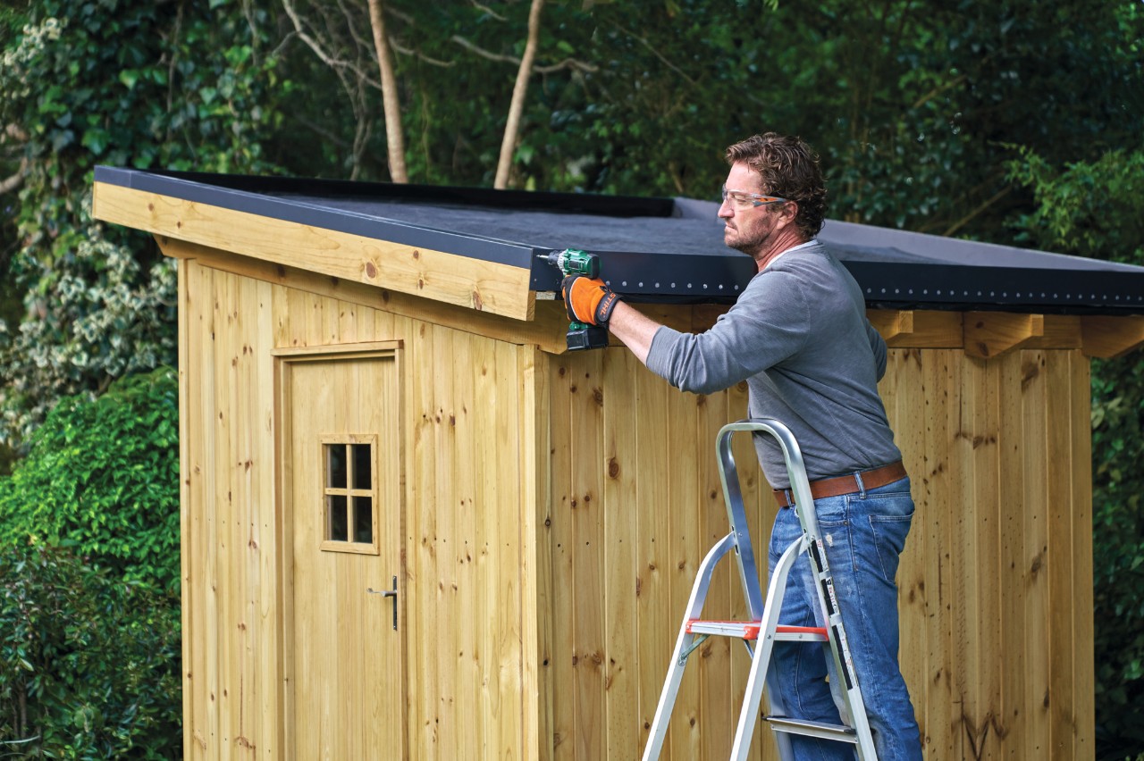 A man on a ladder screwing a green roof retention strip in place on the roof a garden shed