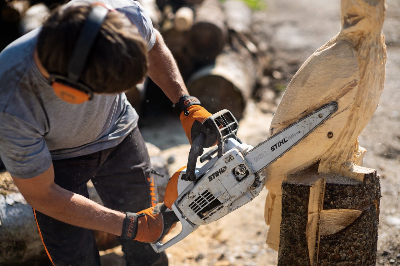 A man wearing ear protection uses a STIHL MS 193 C-E with carving attachment to cut feathers into an eagle sculpture