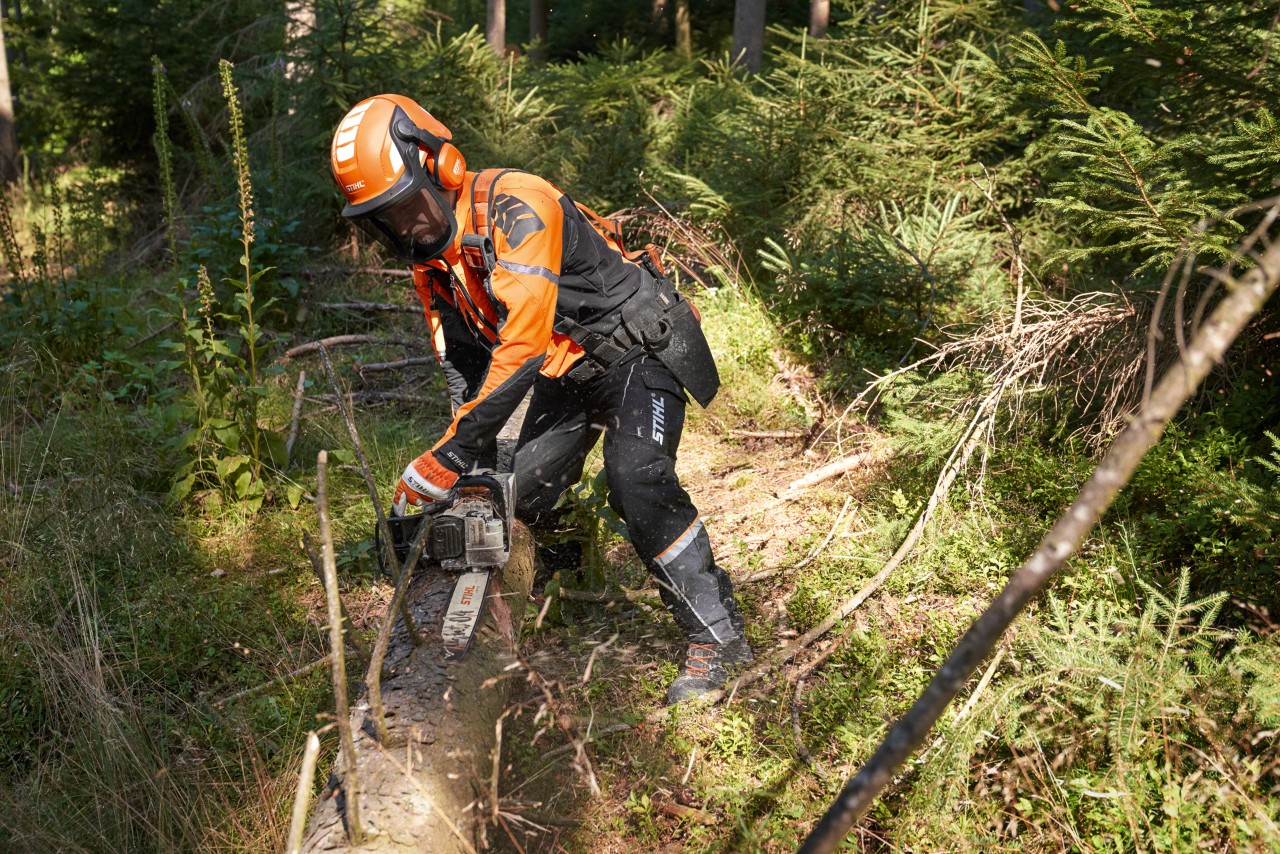 A person wearing protective equipment uses a STIHL chainsaw to remove branches off a tree trunk on the ground