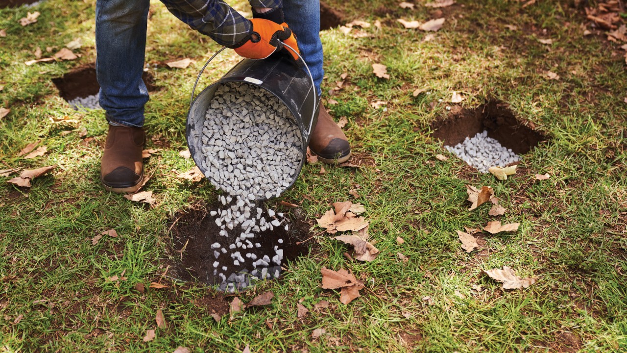 Gravel being poured from a bucket into a square foundation hole in a lawn