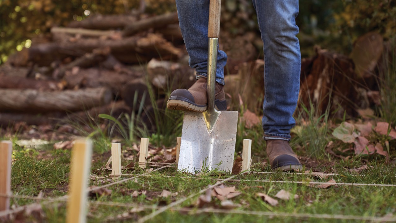 Close-up of a spade being driven into the ground, making a hole in a spot marked by stakes and string