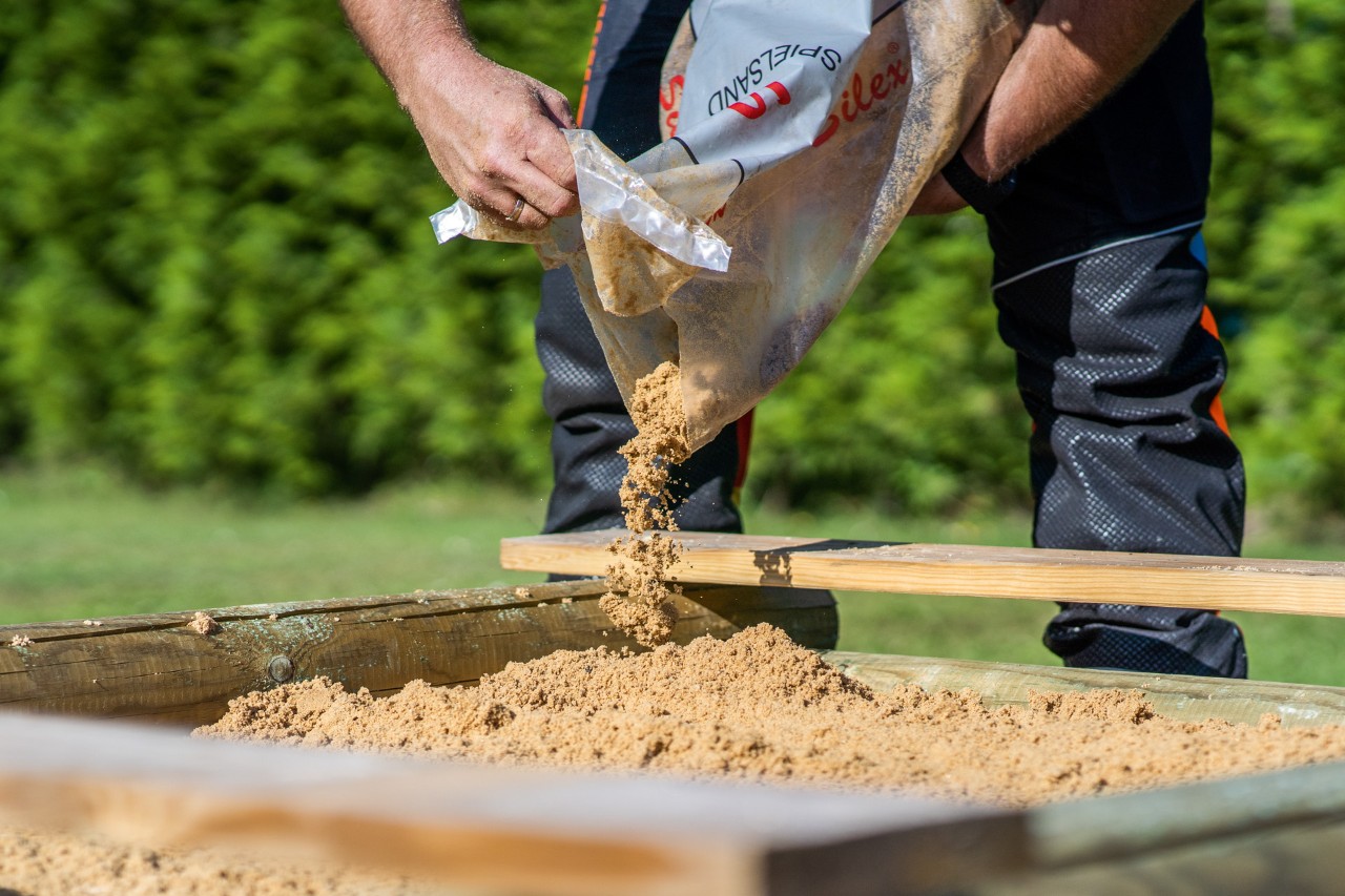 A finished wooden sandpit being filled with sand from a bag.