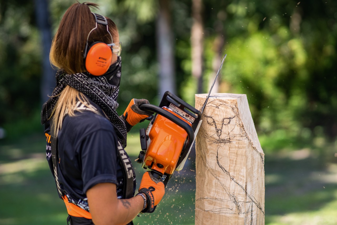 A woman wearing protective equipment uses a STIHL chainsaw to roughly carve a piece of wood with an owl drawn on it.
