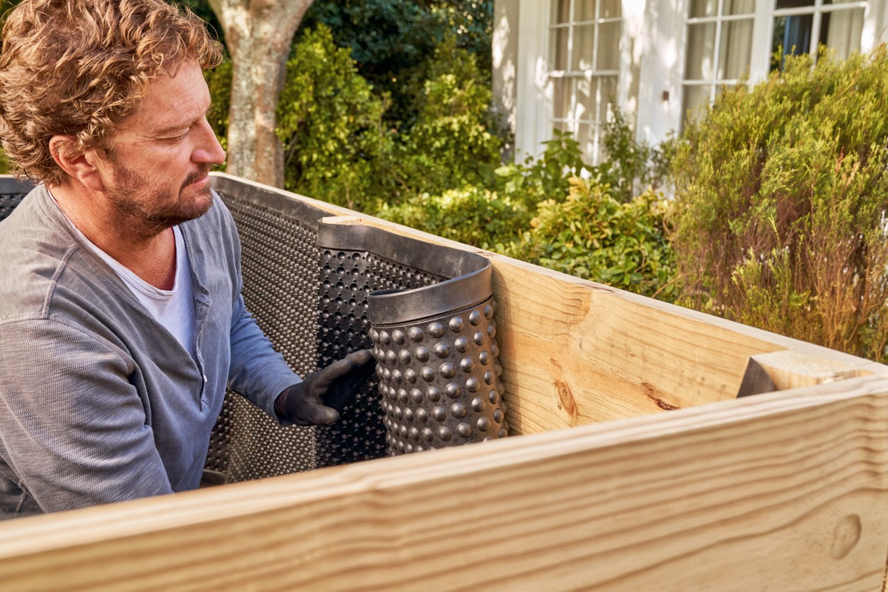 Build a raised bed from boards: a man attaches black waterproof membrane to the inside of a raised bed