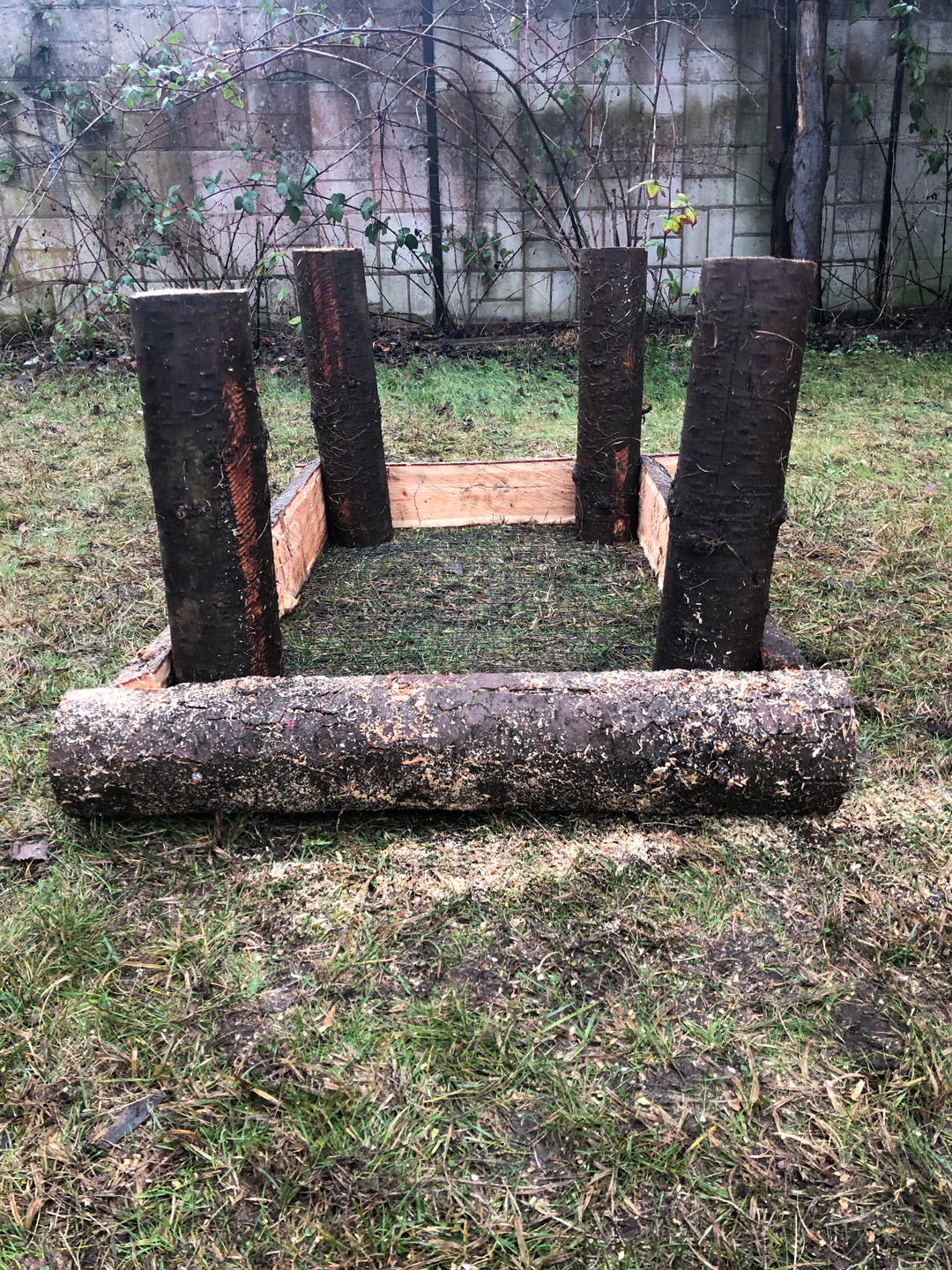 The foundation of a raised wooden bed, with four upright pillars in a rectangle and a row of split logs to secure them