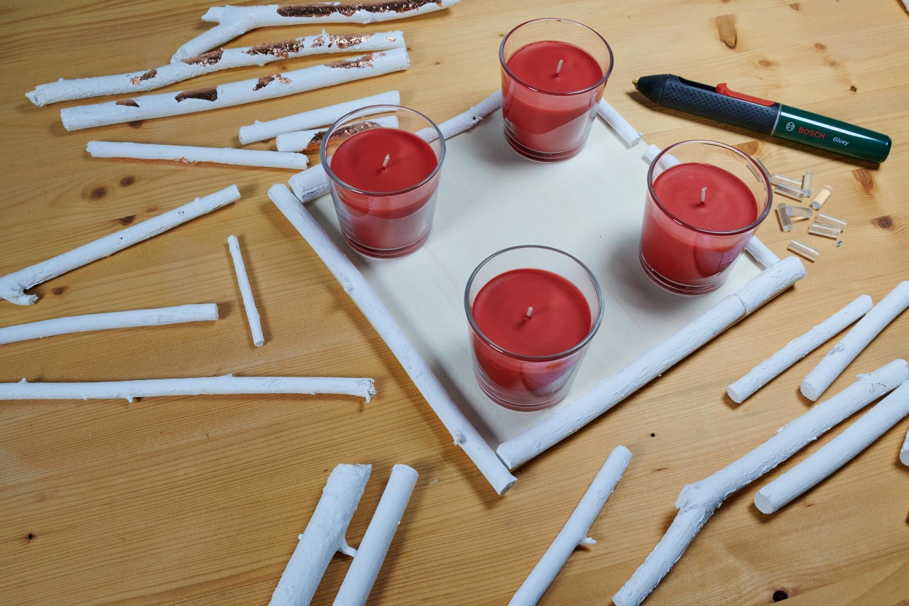DIY advent wreath: four red candles in glasses on a square wooden board, surrounded by white twigs