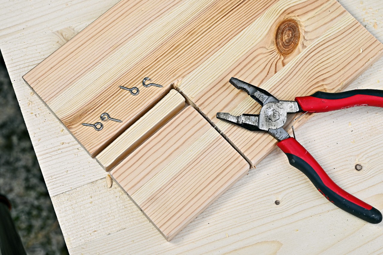 Four screw eyes and a pair of pliers on a wooden board that has been cut to make a flap