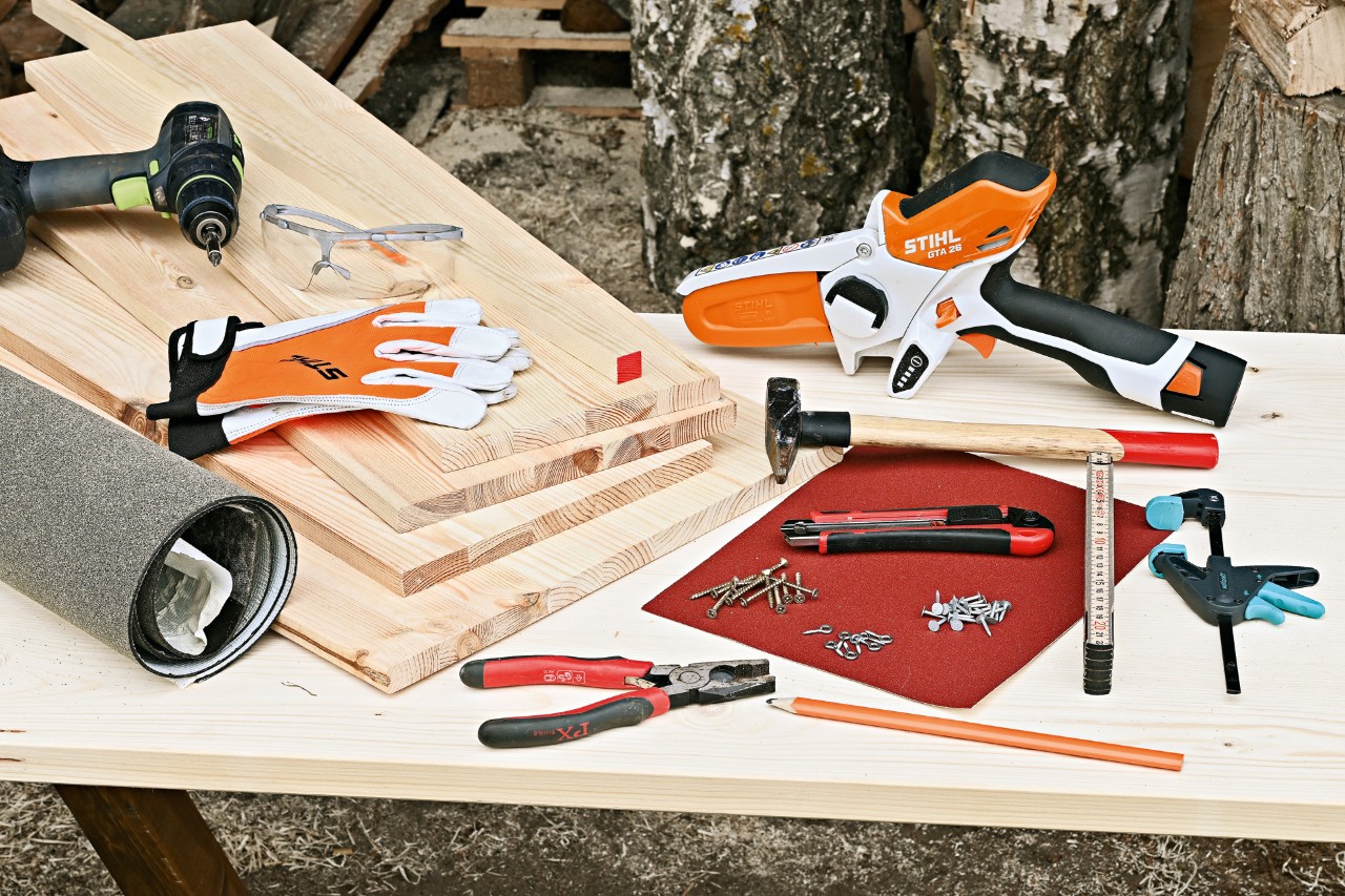DIY hedgehog house tools and materials: STIHL GTA 26 cordless garden pruner, wood panels, hammer, nails, and other tools