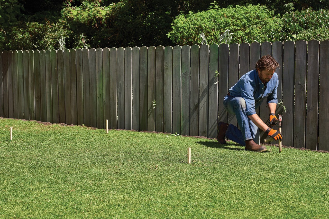 A man staking out the outlines of a vegetable patch on a lawn beside a wooden fence