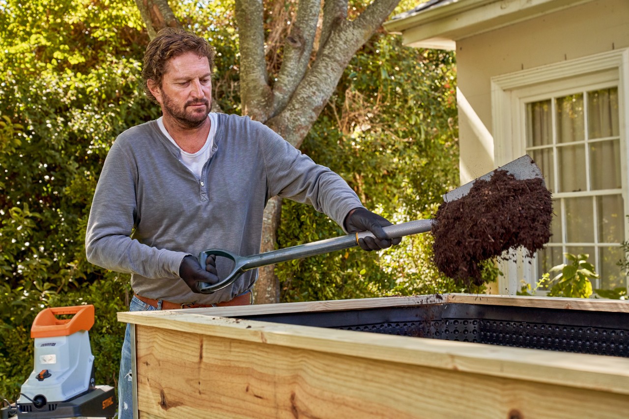 A man using a spade to fill a raised bed with half-finished compost