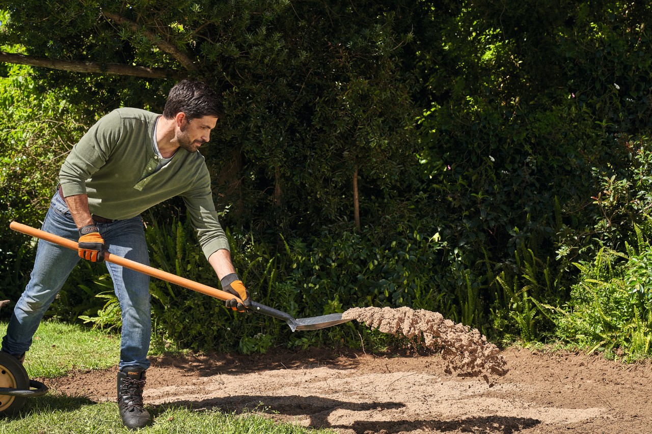 A man spreading building sand on loamy garden soil with a spade to reduce the nutrient concentration  