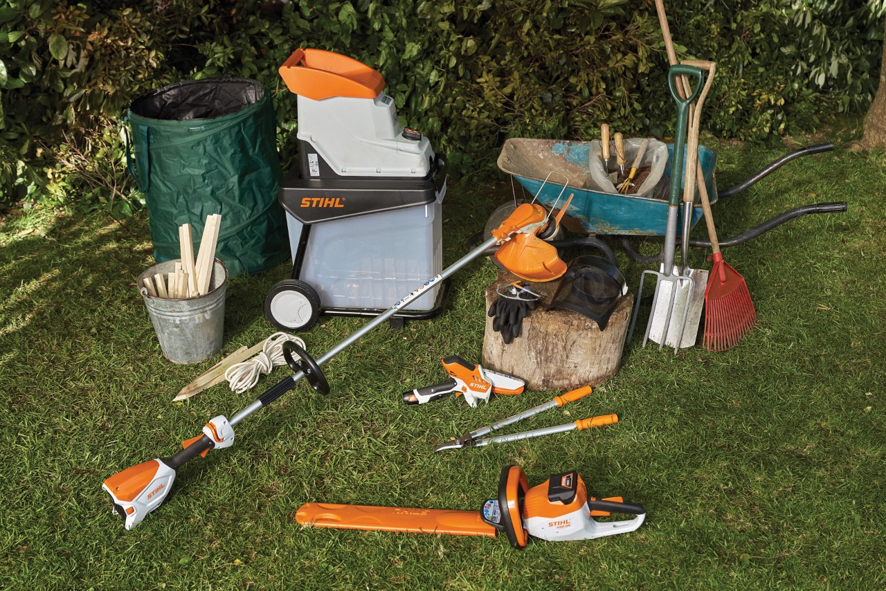A hedge trimmer, garden shredder, wheelbarrow and other tools and equipment laid out on a lawn