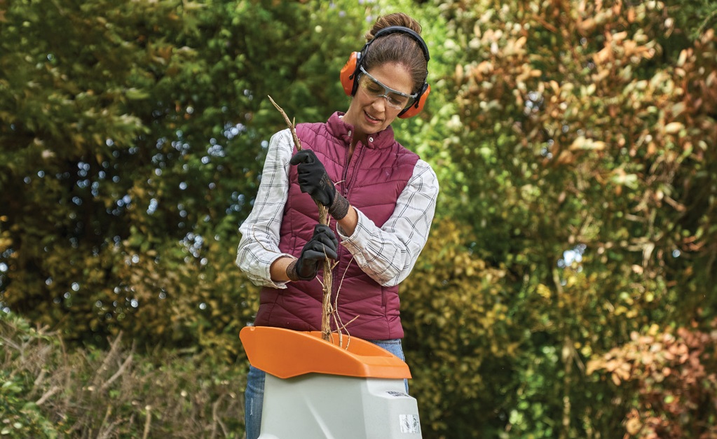 A woman wearing gloves, ear protection and glasses feeding branches into a garden shredder