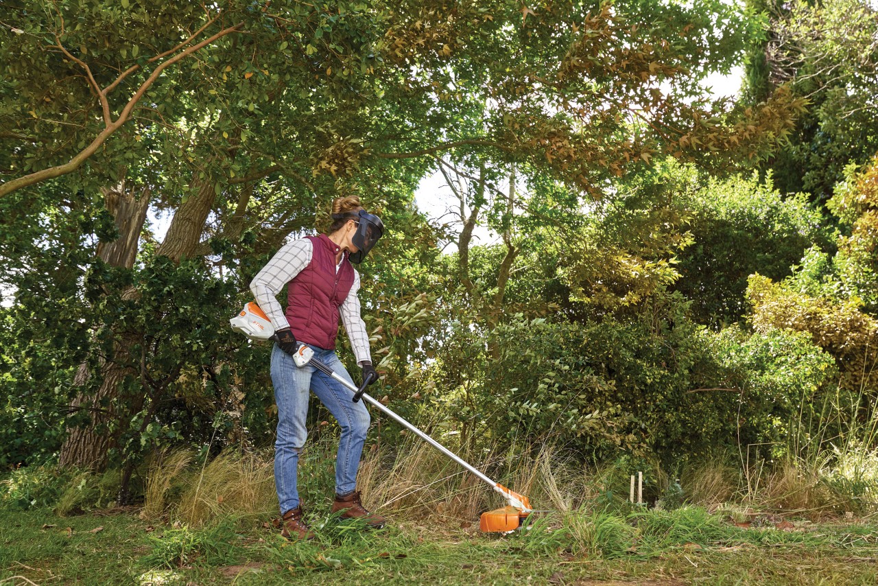 A woman using a brush cutter on overgrown grass with trees and shrubs in the background