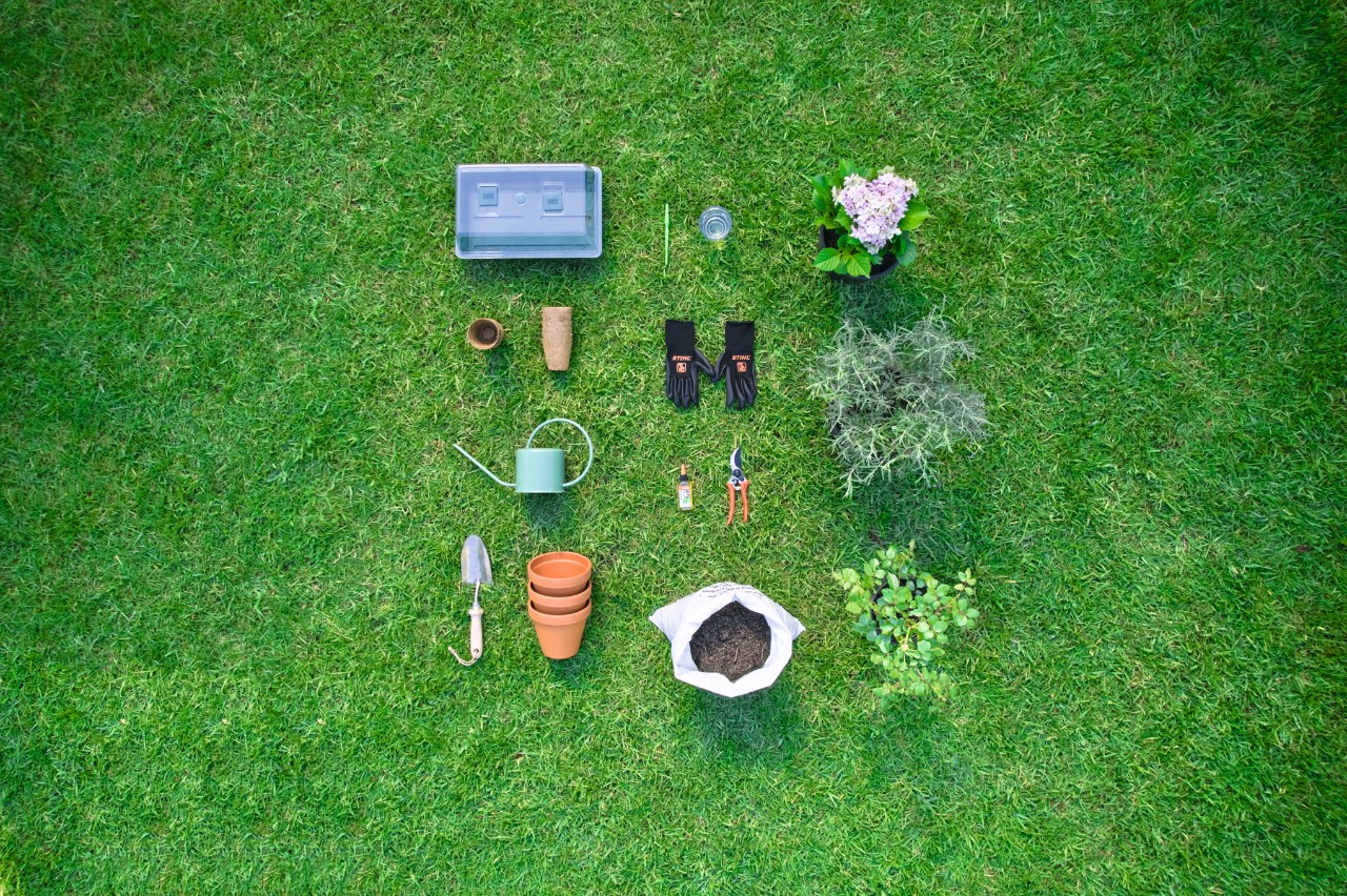 Tools and materials for taking and propagating cuttings laid out on grass, including secateurs, gloves, soil, a spade, pots and a watering can