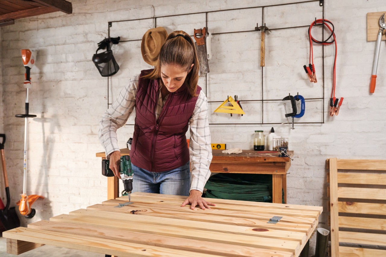 A woman using a cordless screwdriver to screw an access hatch onto the front of the compost bin on a workbench