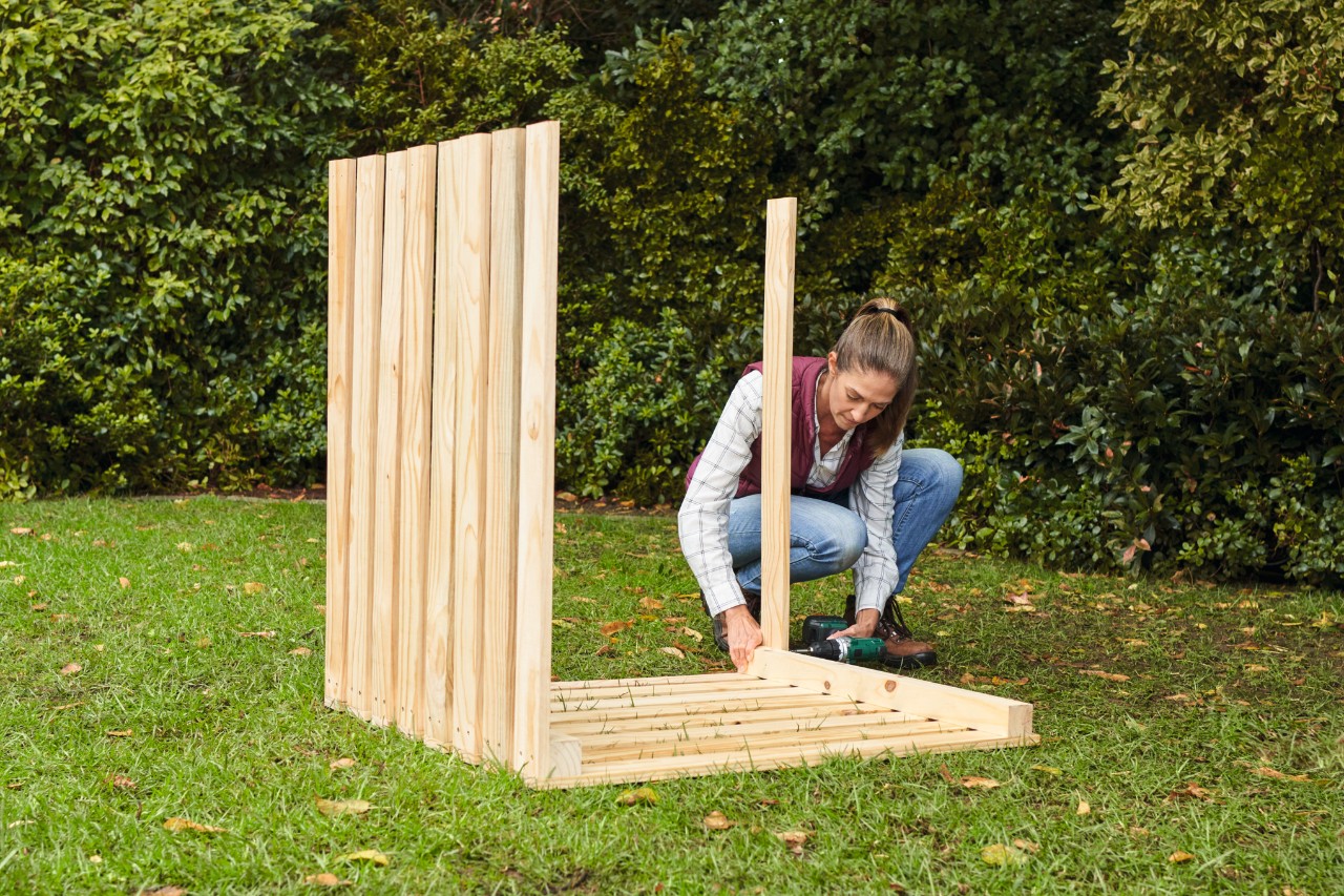 A woman wearing jeans and a ponytail using a cordless screwdriver to attach the slats for the side walls to the composter on a garden plot