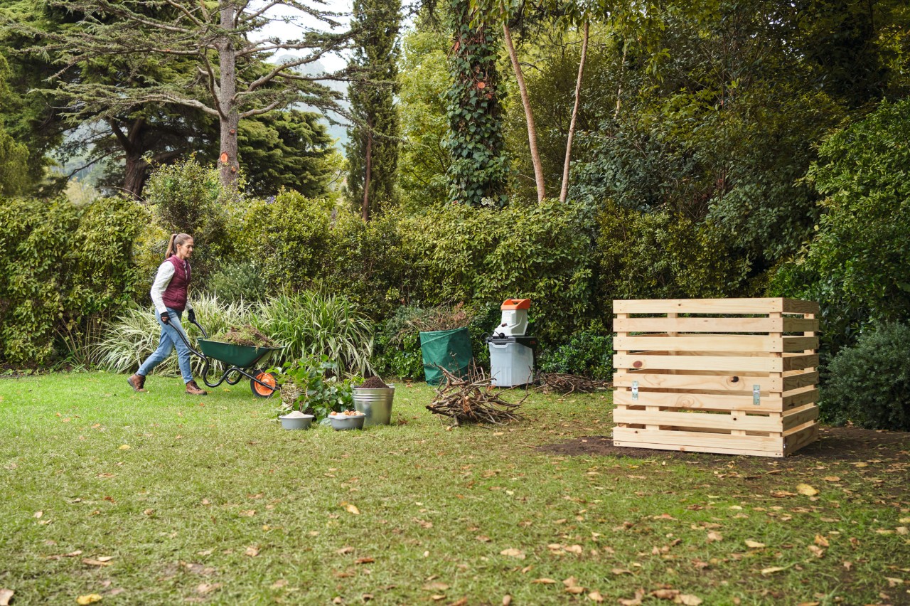 A woman approaching a DIY wooden compost bin with a wheelbarrow, with a dense hedge and trees in the background 