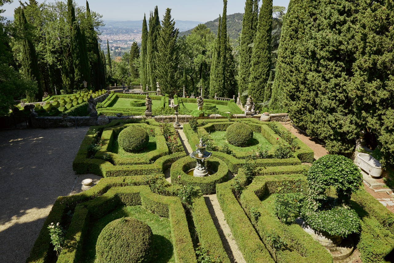 View over a formal garden containing geometric hedging borders and topiary balls, with tall trees in the background.