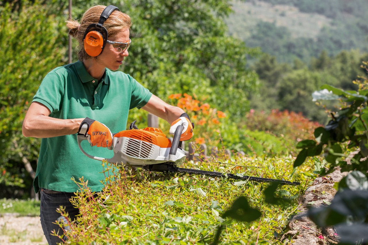 A woman with hearing protection and protective goggles cuts a hedge with a STIHL HS 46 petrol hedge trimmer