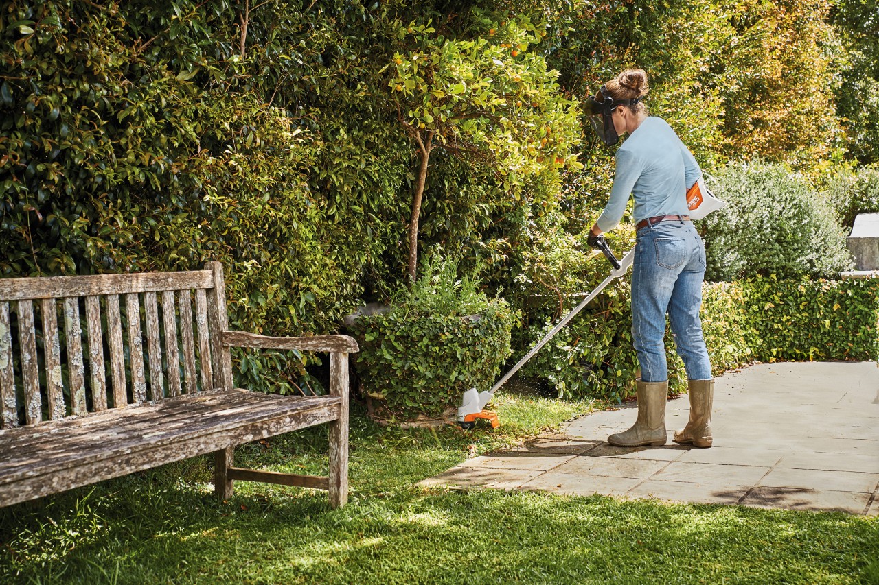 A woman trimming the edge of a lawn around some bushes with a STIHL FSA 57 cordless clearing saw