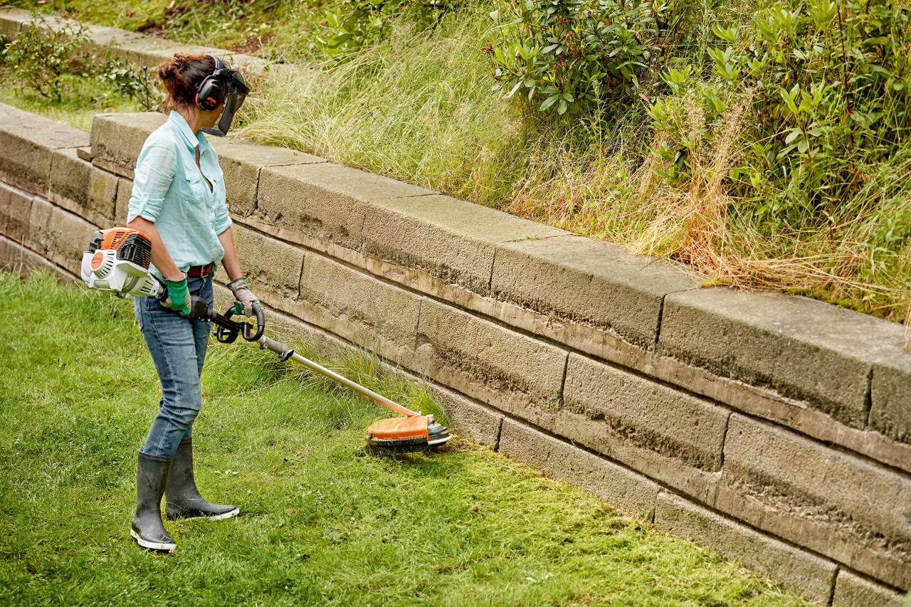 A woman wearing ear and face protection using a STIHL FS clearing saw to trim the edge of a lawn along a wall
