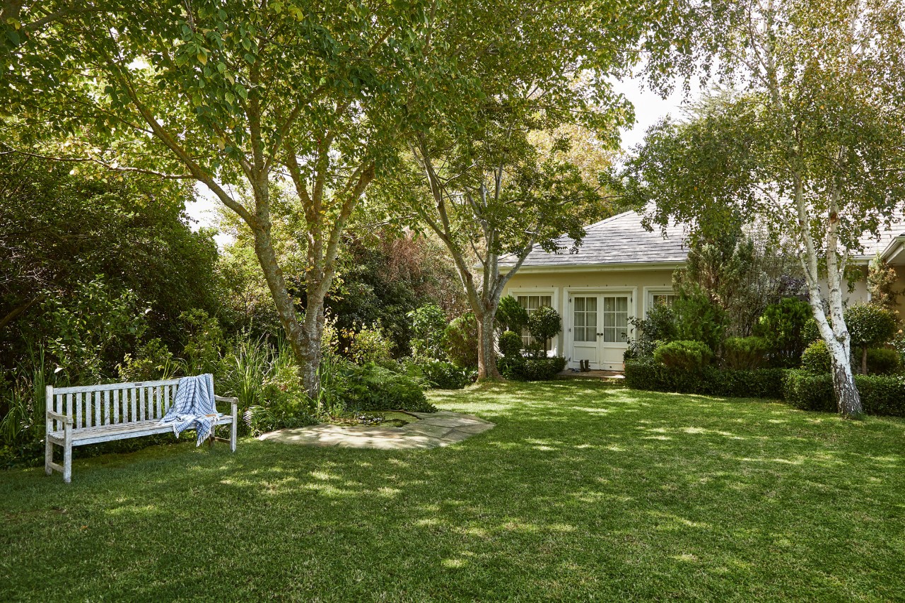 A plot with trees and a garden bench on a shaded lawn in front of a house.