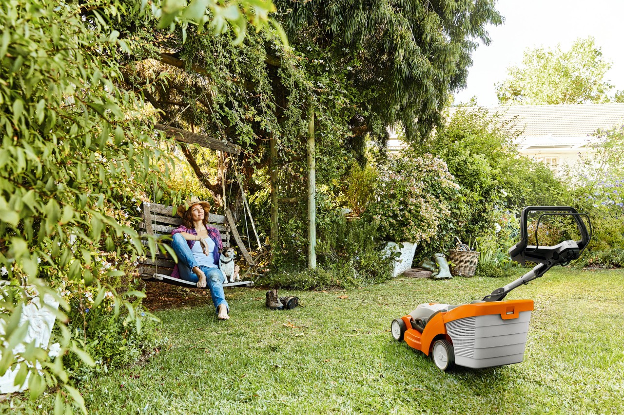 A woman relaxes on a garden swing next to a STIHL RMA 339 cordless lawnmower on a green plot