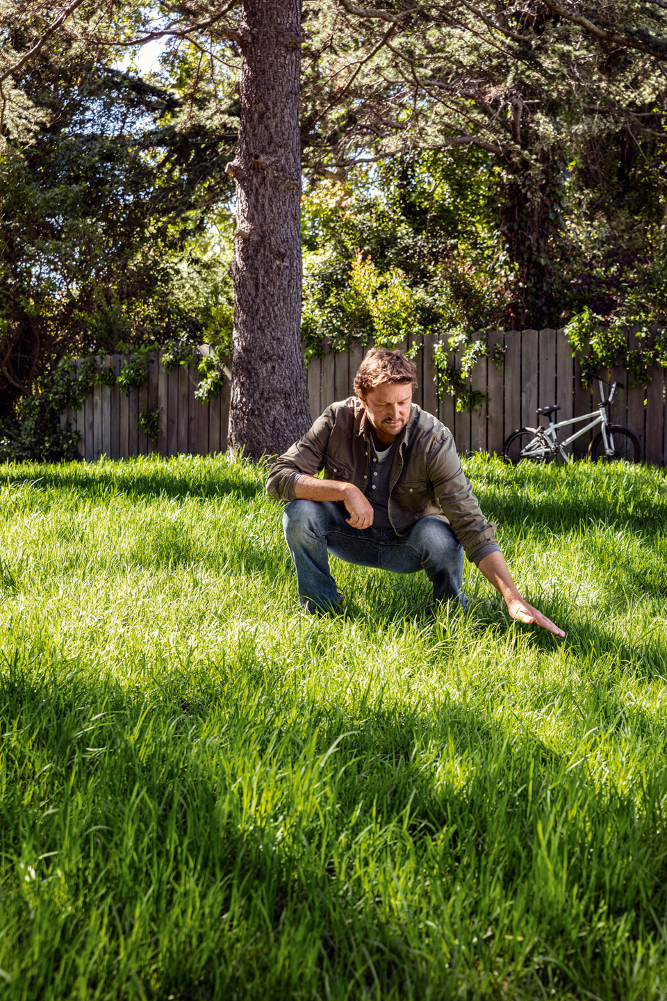 A man crouches down on a lush, healthy green lawn, looking at it and touching it with one hand. There is a tree and a bike in the background.