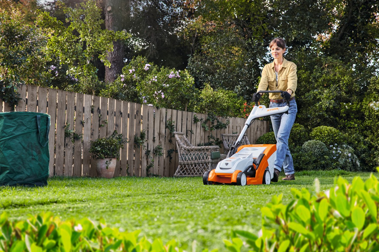 A man is halfway through mowing untidy and overgrown grass using a STIHL RMA 448 P-C cordless lawn mower, in a garden surrounded by tall shrubs and plants