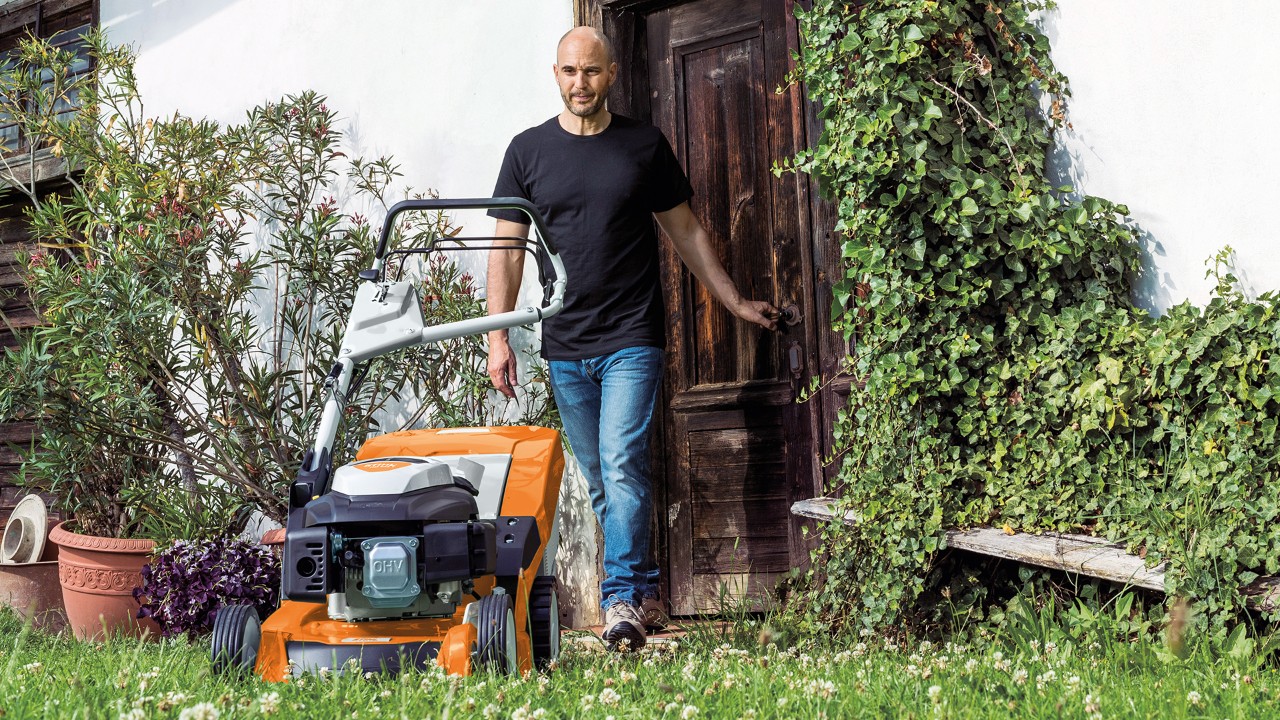 An unmown lawn outside a white building, from which a man walks towards a STIHL RM 650 V petrol mulching mower