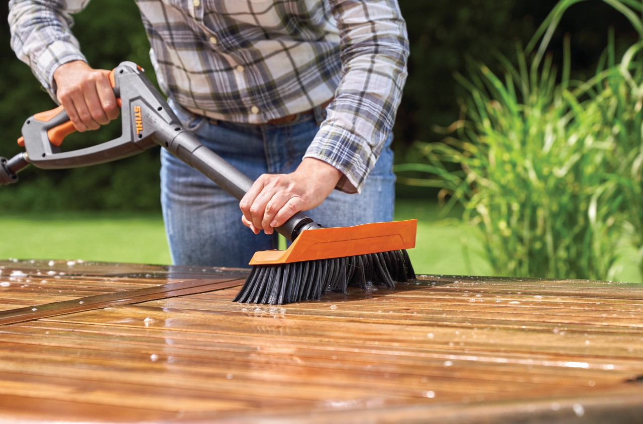 A woman cleaning a wooden garden table using a STIHL RE 130 Plus high-pressure cleaner with wash brush