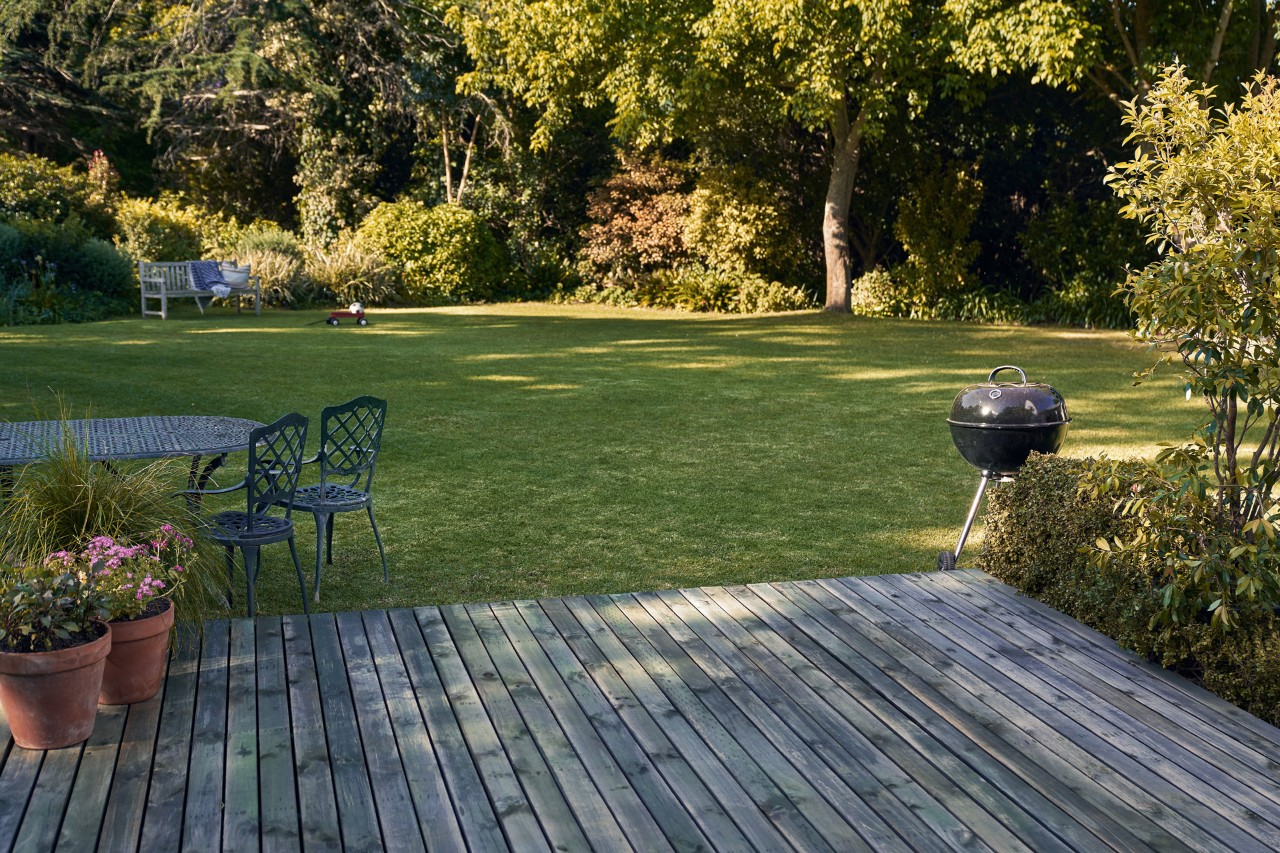 Wooden terrace in front of a green garden with barbecue, chairs and flower pots
