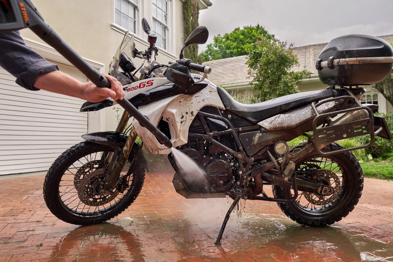 A motorcycle on a driveway in front of a white house, being cleaned with a STIHL RE 130 plus pressure washer