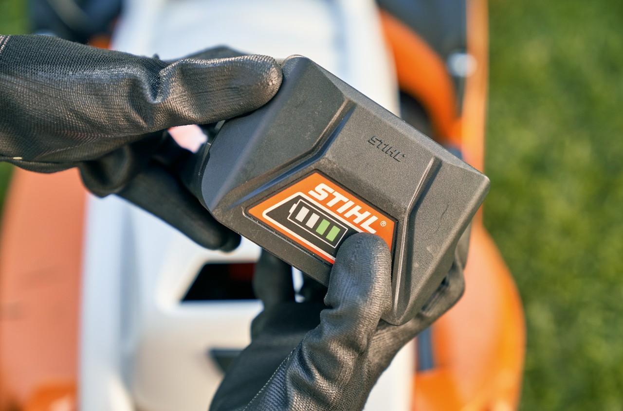 A STIHL Li-ion battery being held in gloved hands above a STIHL RMA 339 C cordless lawn mower