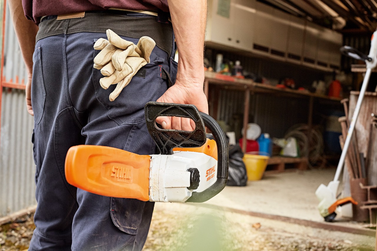 A man walking into a workshop holding a STIHL cordless chainsaw with scabbard