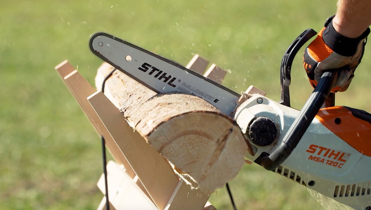 A log on a sawhorse is cut into firewood using a STIHL chainsaw MSA 120 C by someone wearing hand protection.