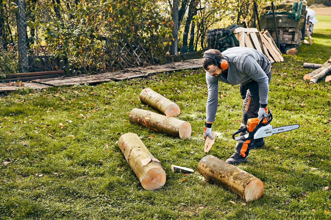 A metre-long log is secured with wedges by someone wearing PPE and holding a STIHL MS 180 petrol-driven chainsaw.
