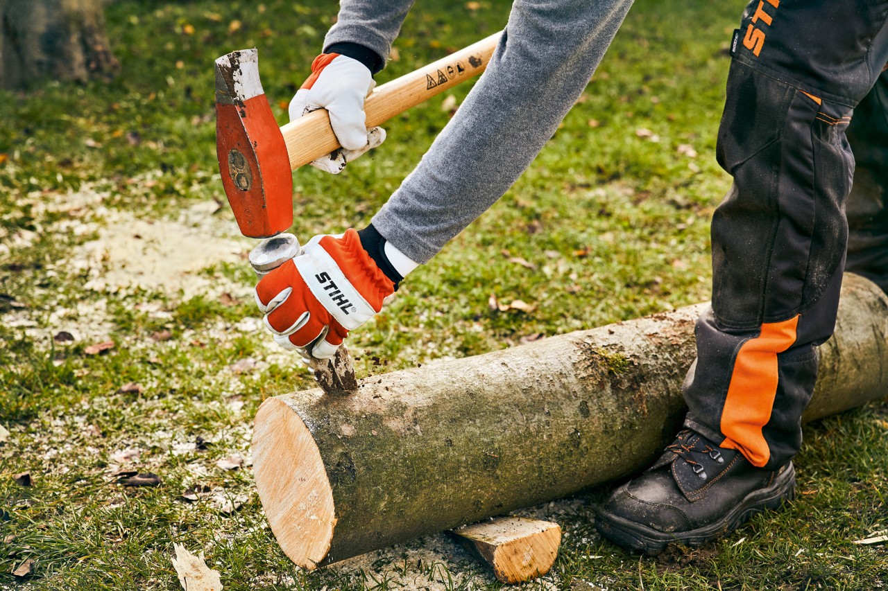 Someone wearing gloves using a splitting wedge and hammer on a length of trunk