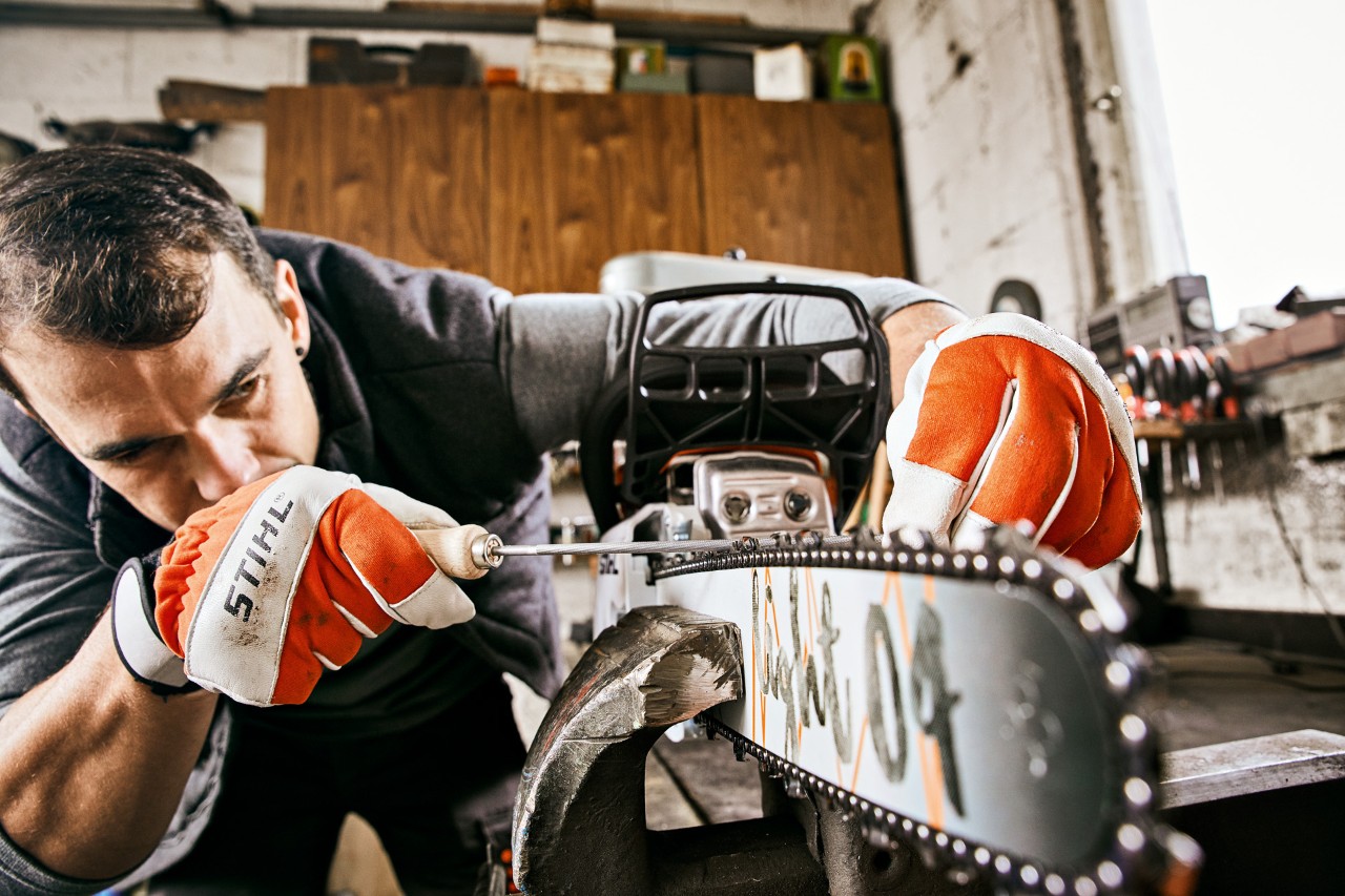 A STIHL 261 C-M petrol chainsaw clamped to a workbench as a man wearing gloves uses a round file to sharpen the chain