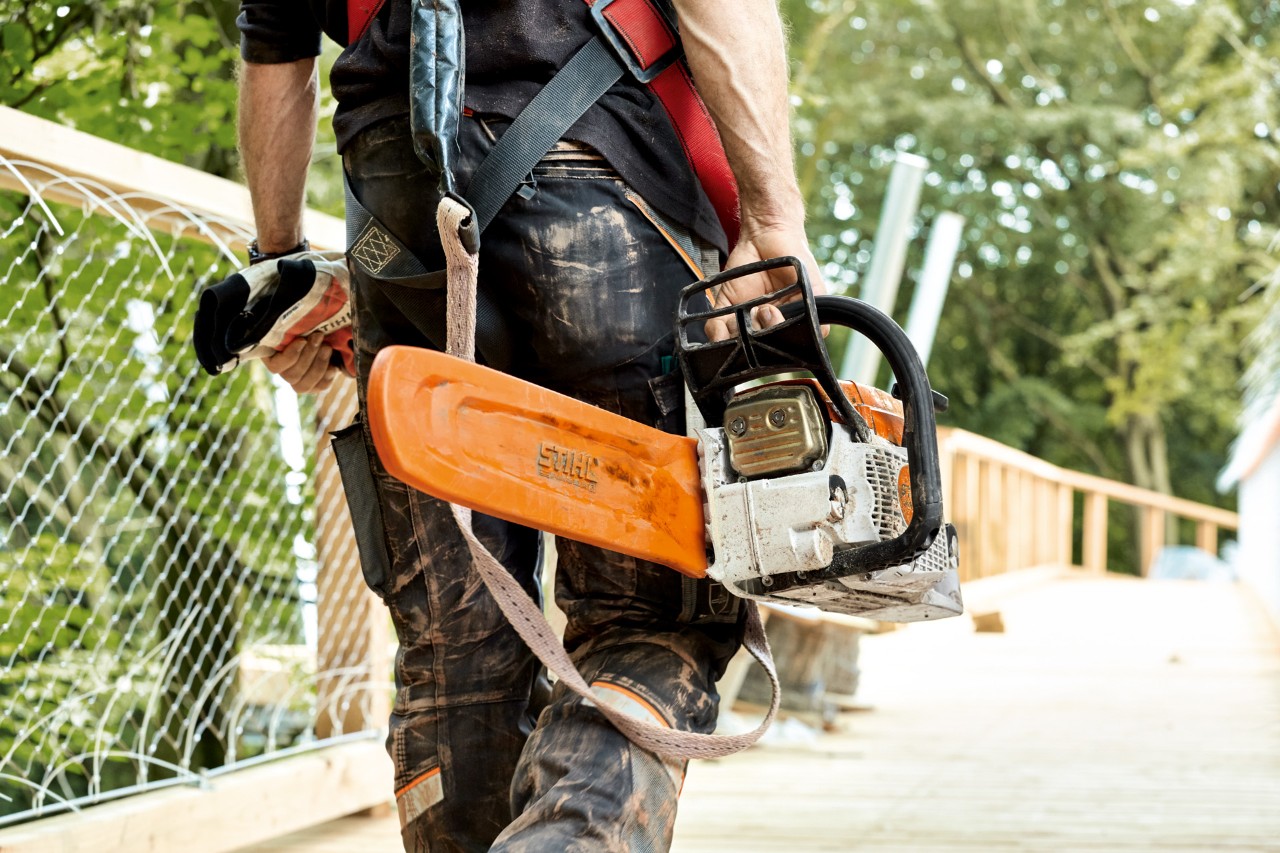 A STIHL MS 261 petrol chainsaw with chain guard being carried by a man as he walks across a bridge