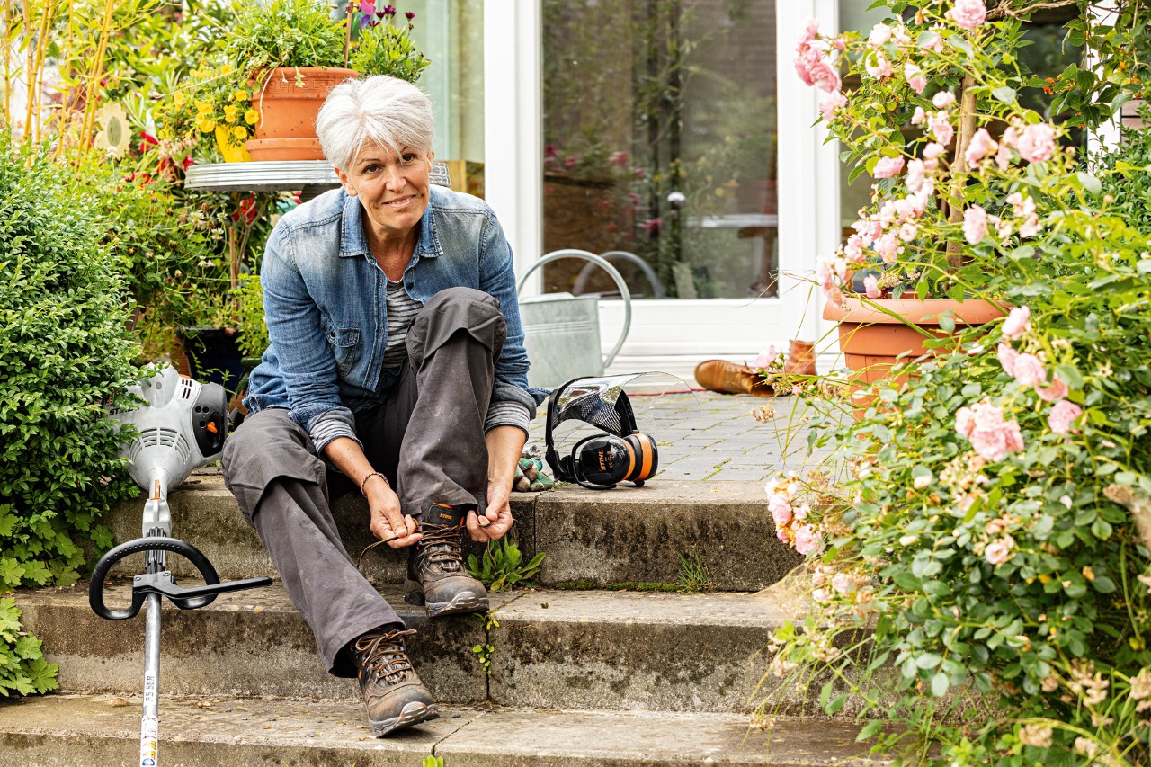 A woman sitting on steps tying a bootlace with a STIHL FS 58 R petrol grass trimmer and visor with ear defenders nearby