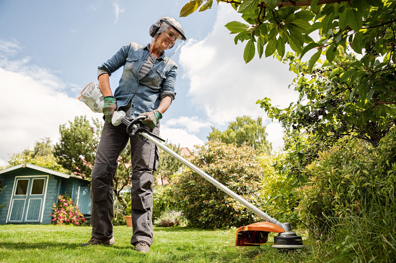 A person wearing a face visor and ear protectors, using a STIHL FS 58 R petrol grass trimmer at the edge of a lawn