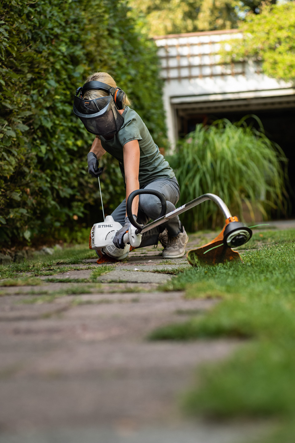 A woman wearing protective equipment and gloves starts a STIHL FS 38 petrol grass trimmer that is on a path in a garden