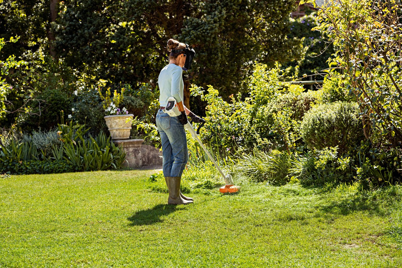 A woman trims tall grass around the edge of a garden using the STIHL FSA 57 cordless grass trimmer