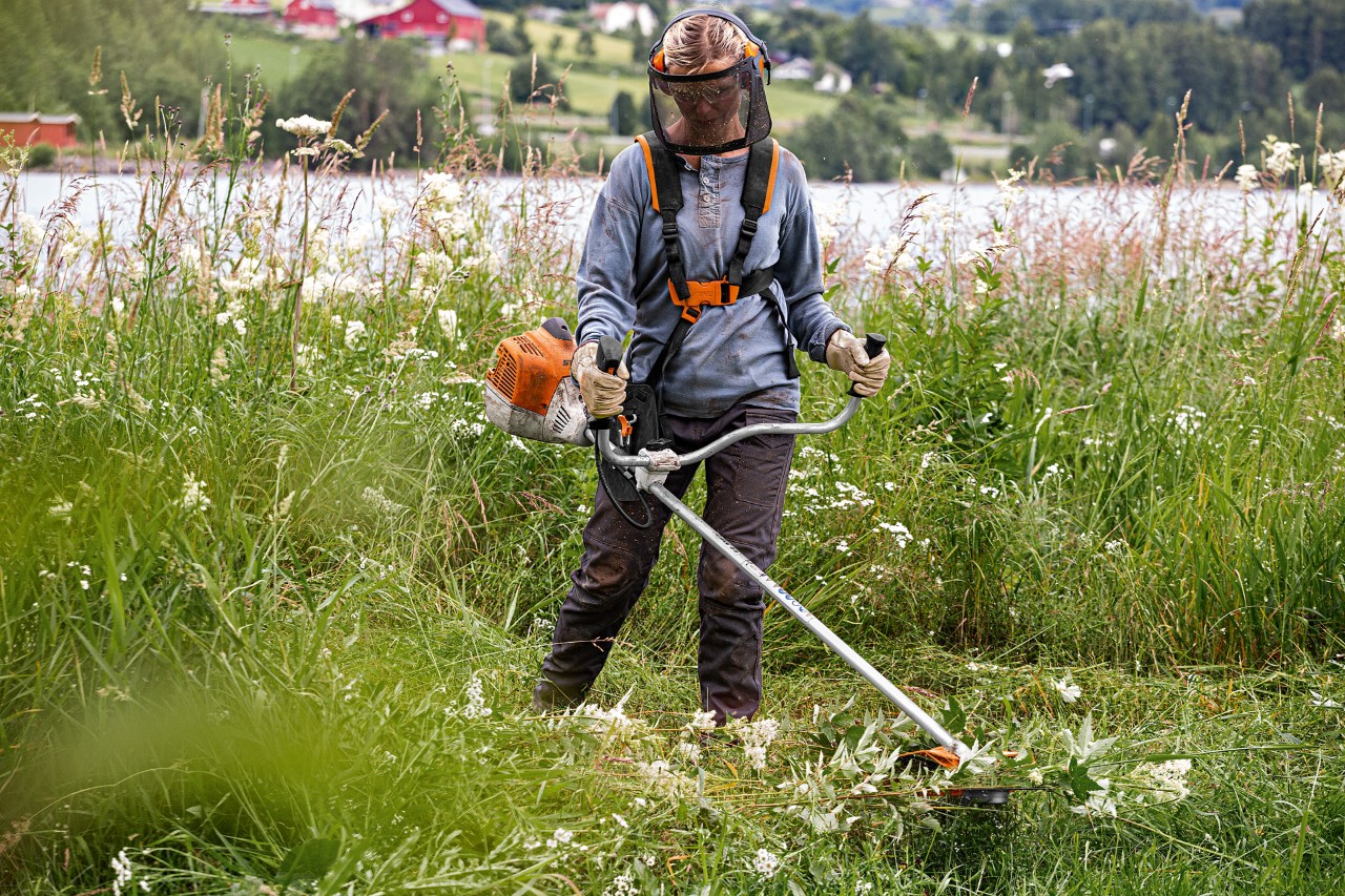A woman wearing face protection uses a STIHL FS 240 grass trimmer in long grass beside a lake
