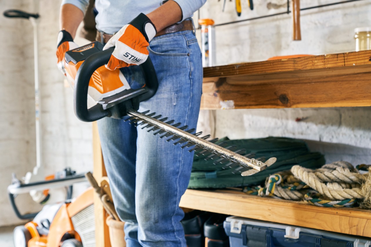 A STIHL HSA 56 cordless hedge trimmer being held by someone wearing gloves, in front of a workbench and lawn mower
